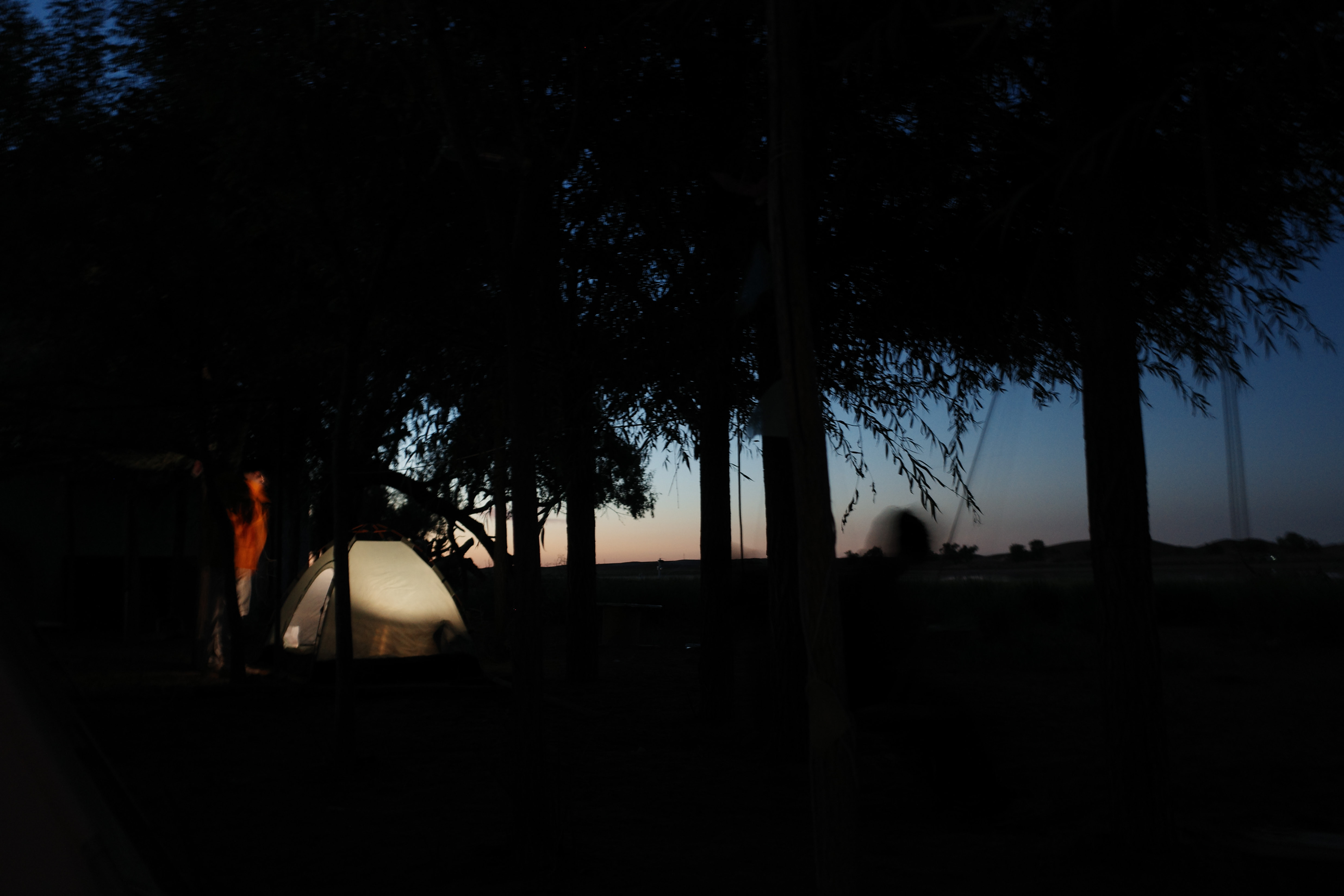A lit tent glowing under trees at night with a silhouetted figure in orange nearby