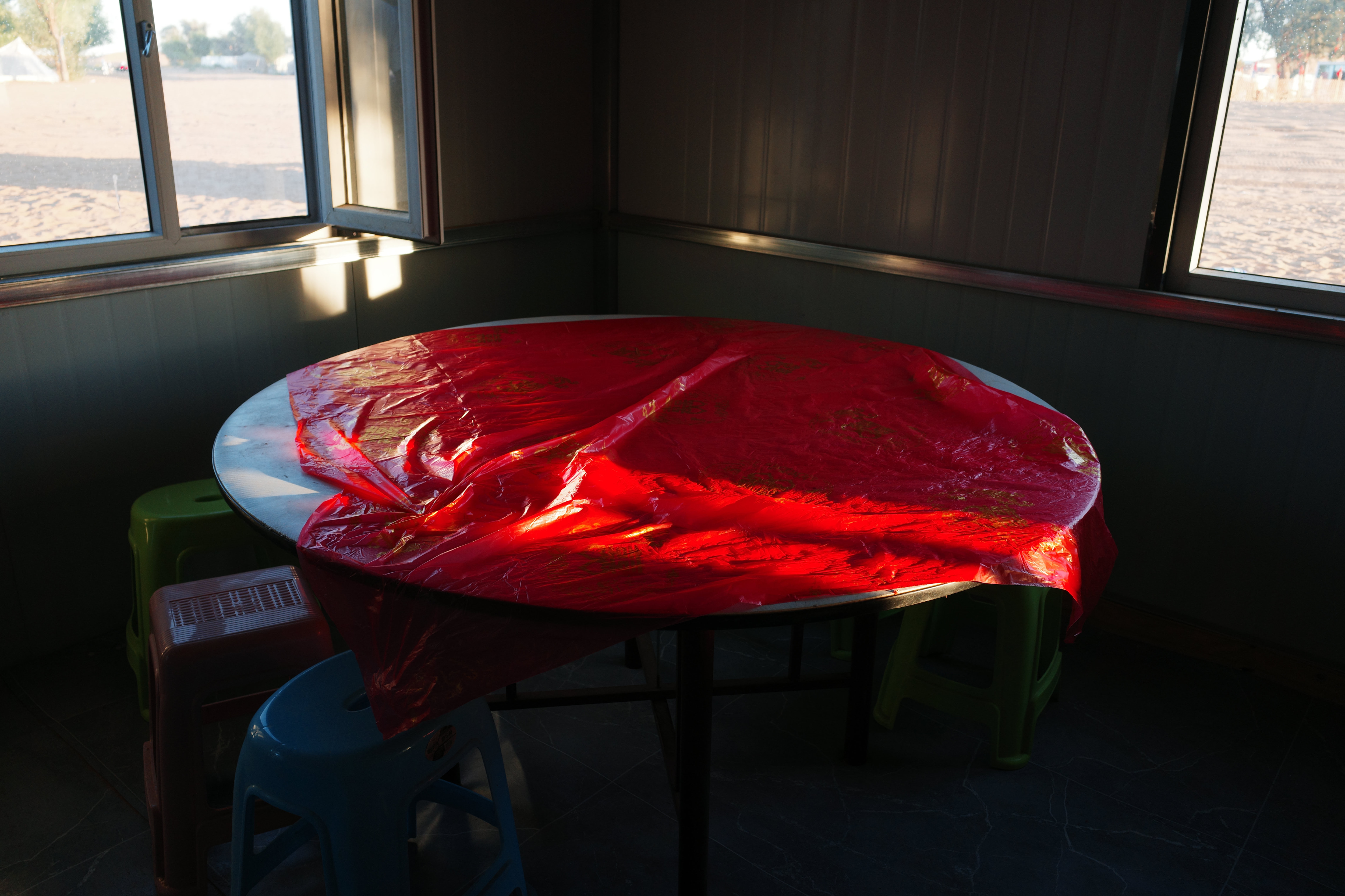 A round table under a wrinkled red plastic cloth catching low sunlight, plastic stools around it