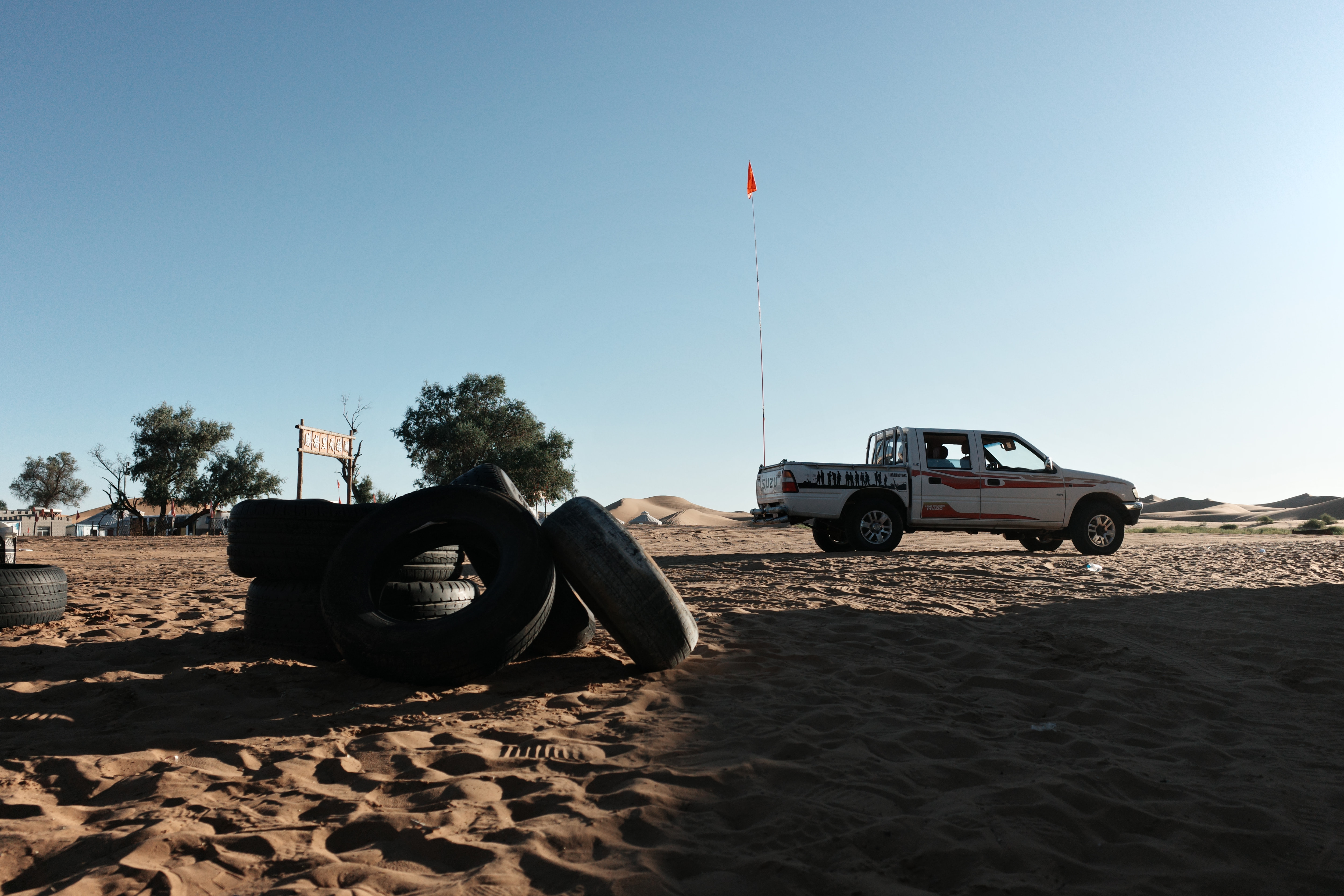 Stacked tires in the sand beside an off-road pickup truck flying a red flag, dunes behind