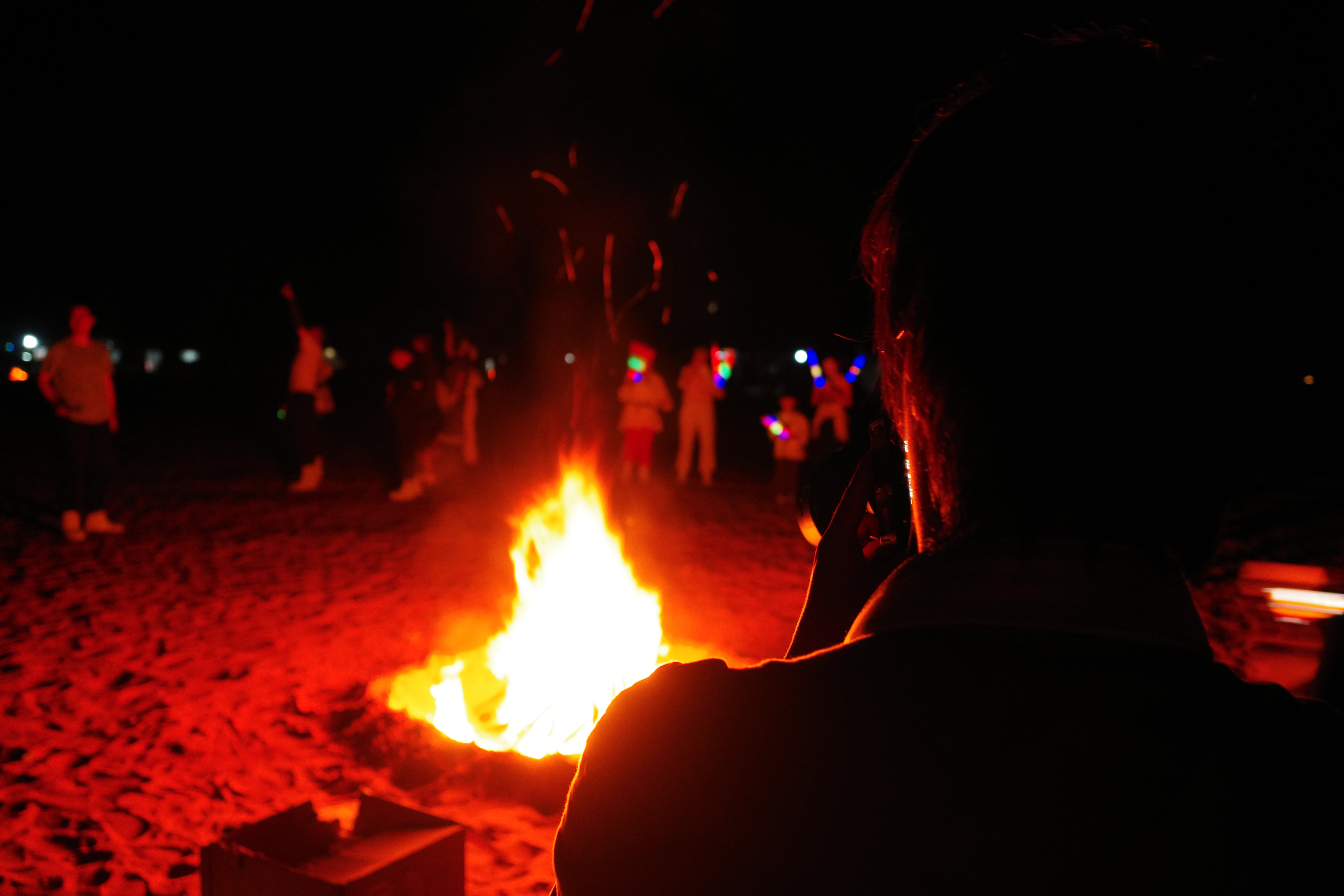 Bonfire at night with onlookers holding colored glow sticks, a figure with a camera in the foreground