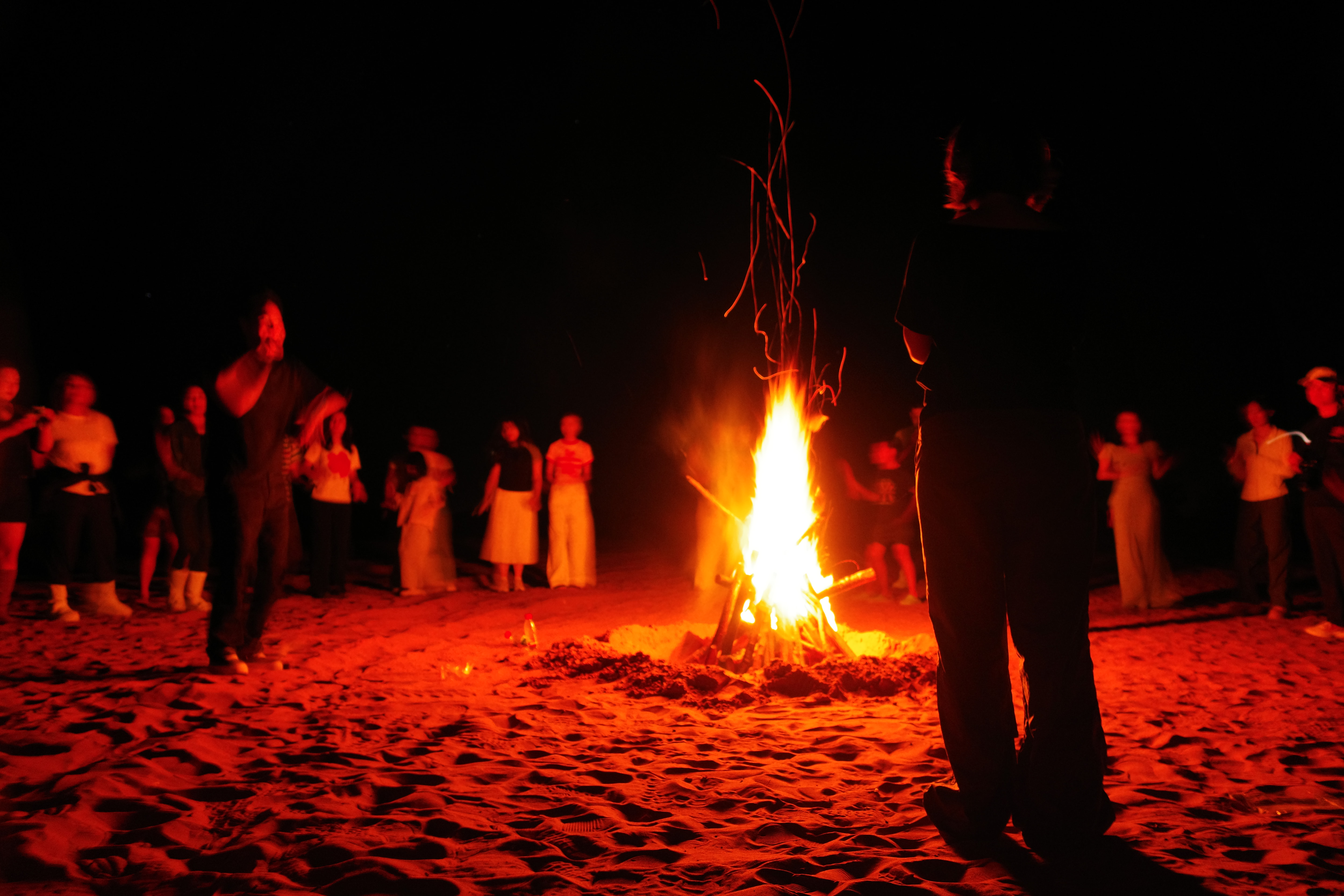 Long-exposure night photo of a circle of people dancing around a bonfire on sand