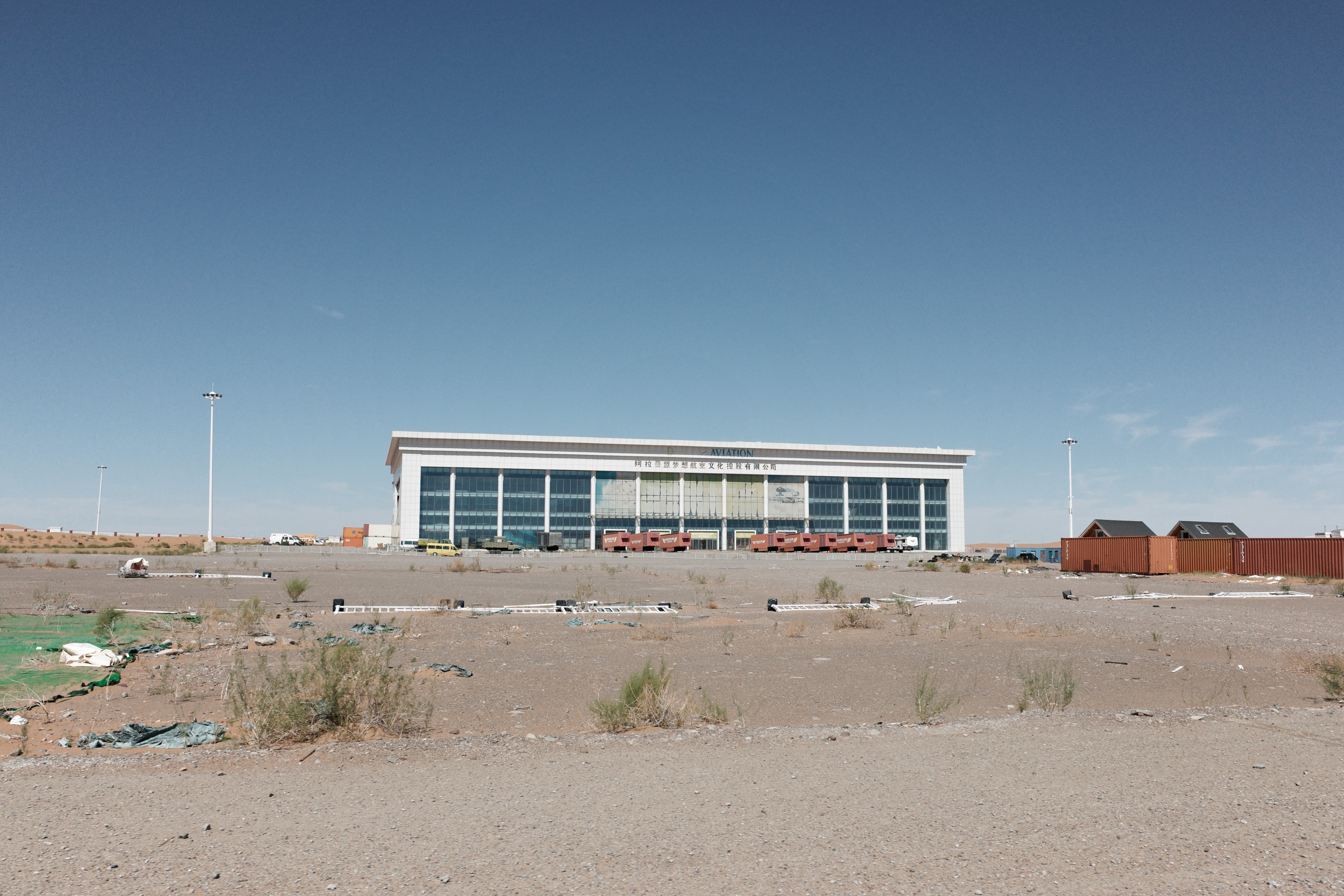 A large glass-fronted building labeled Aviation in the desert, shipping containers parked in front