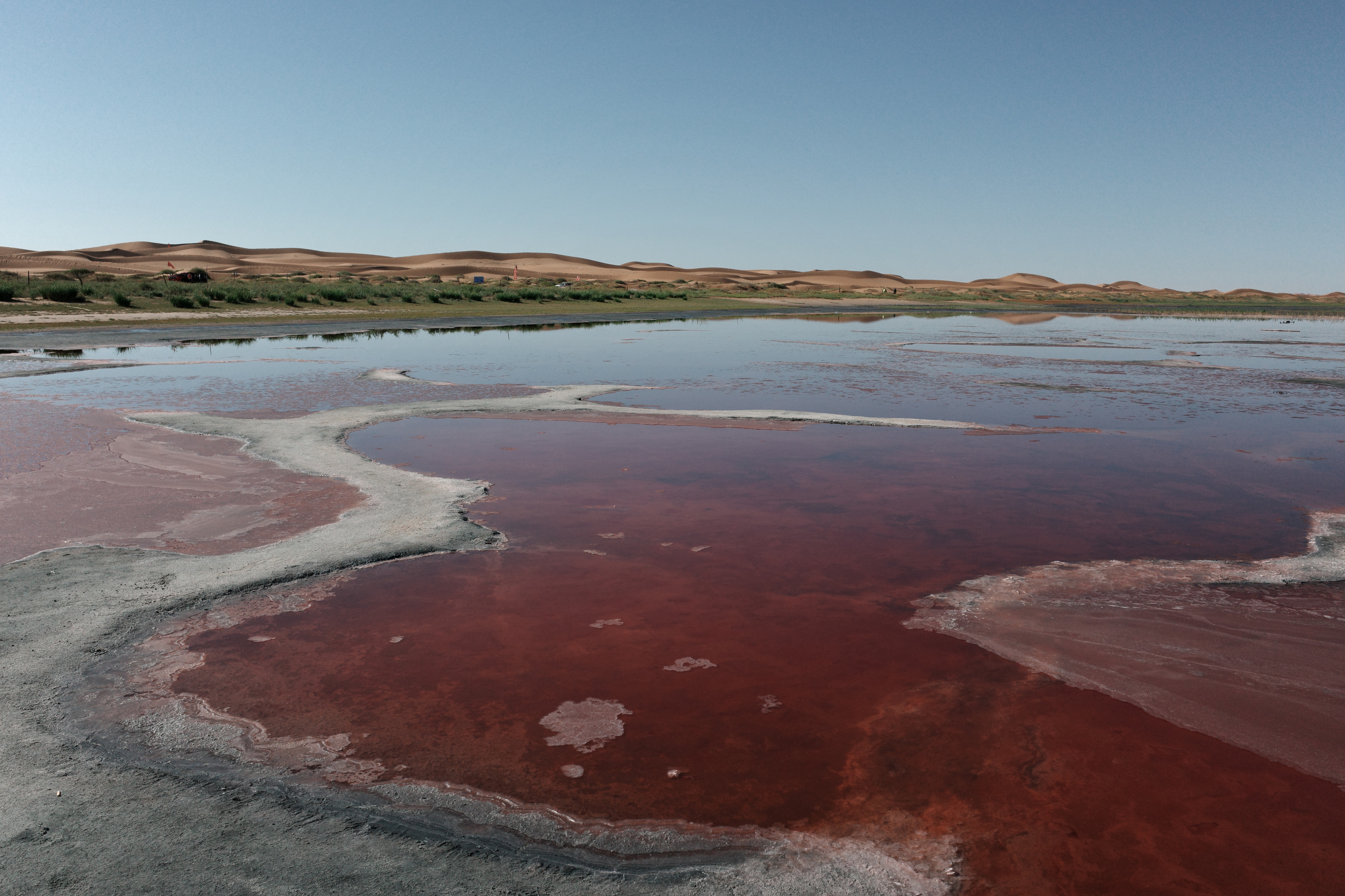 A rust-red saline pool with white mineral crust at its edges, dunes in the distance